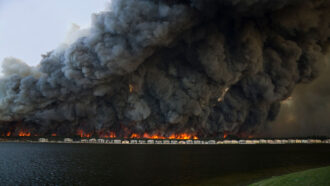 a photo of houses on a lakefront with a huge brushfire burning behind them and creating a massive black cloud