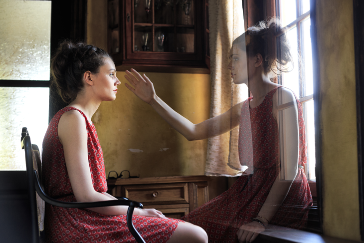 a woman sitting on a chair looking at a ghost twin image sitting in front of a window